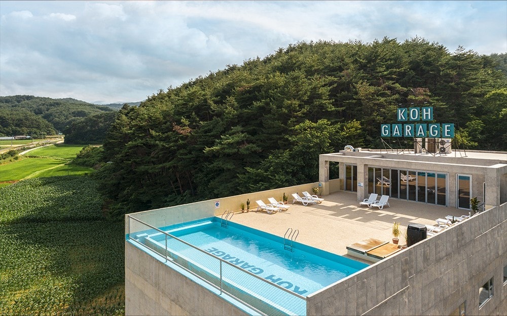 A rooftop area showcases a sparkling outdoor pool with lounge chairs arranged nearby. The space is surrounded by lush greenery, providing a serene backdrop. The 'Koh Garage' sign adds a modern touch, while large windows in the structure allow for ample natural light.