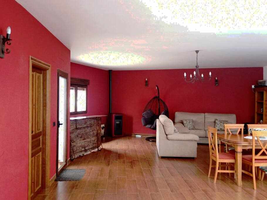 A spacious living area features warm red walls and ample natural light. A comfortable sofa is positioned adjacent to a wooden dining set. A cozy wooden cabinet and a hanging chair enhance the space, while a stove is visible in the corner, providing additional warmth.