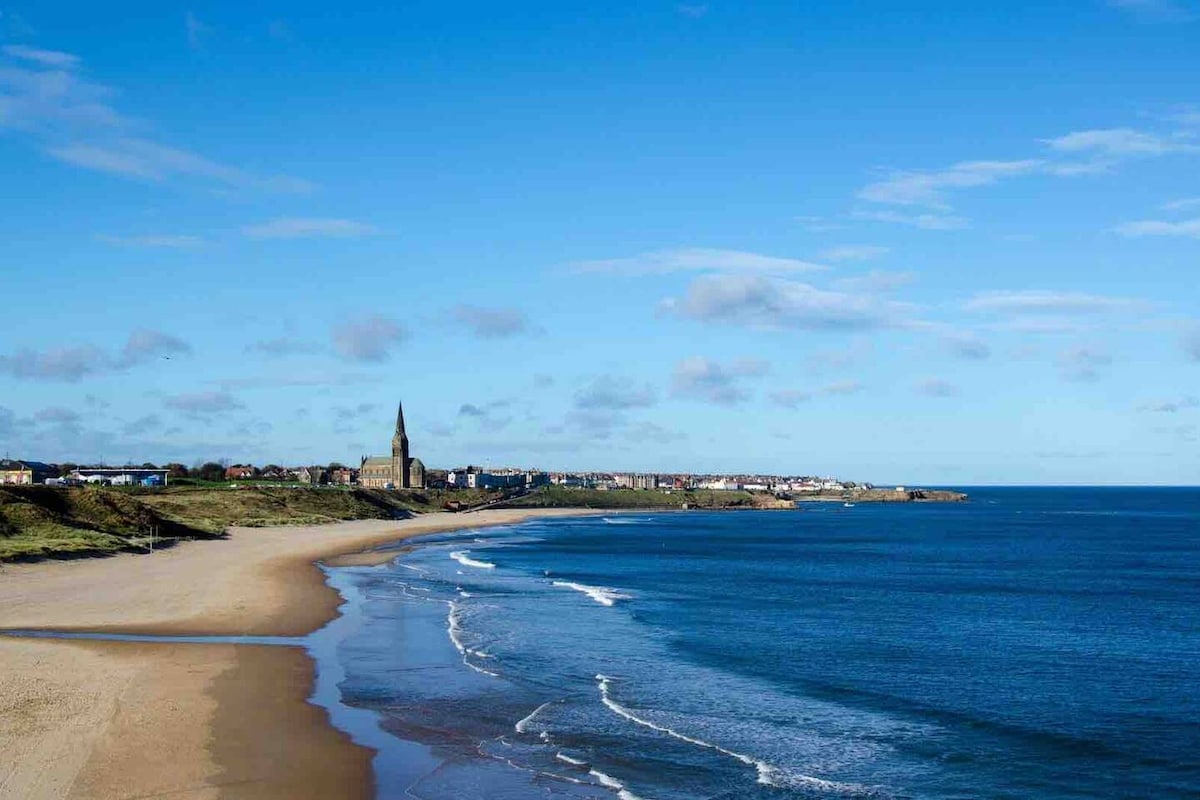 A sandy beach stretches along the coastline, meeting gentle waves lapping at the shore. Scattered clouds drift above, complementing the clear blue sky. In the distance, the silhouette of a village is visible, featuring a prominent church spire and a mix of coastal buildings.