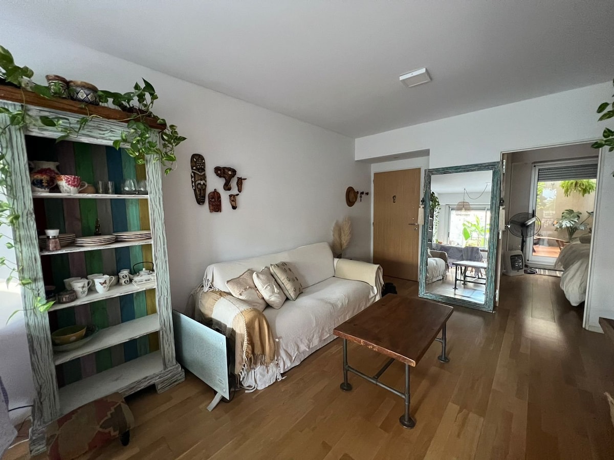 A living area is presented featuring a comfortable sofa adorned with neutral-toned blankets. A wooden coffee table sits centrally, while a decorative shelving unit, filled with pottery and plants, adds character. Natural light flows in through a nearby glass door leading to the terrace.