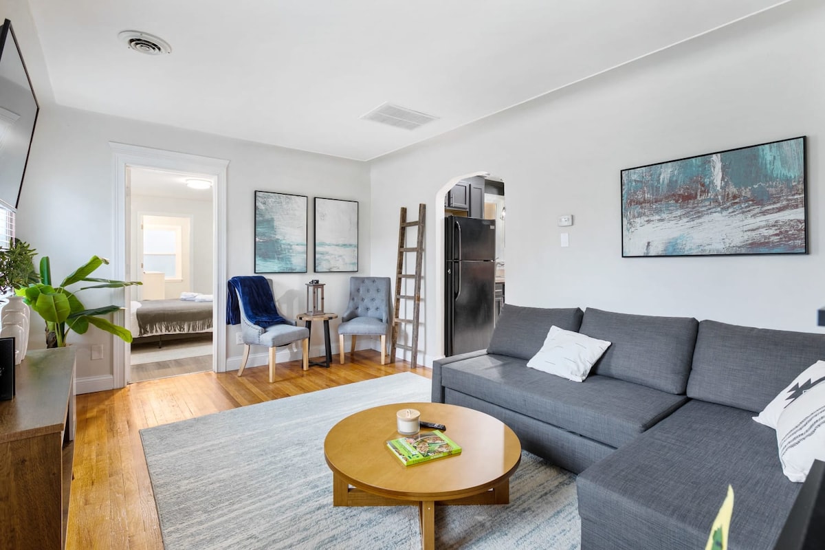 A modern living area features a gray sectional sofa positioned next to a round wooden coffee table. Light hardwood flooring contrasts with the cool tones of the area rug. Two plush chairs add seating, while art pieces adorn the walls, and a hallway leads to additional spaces.