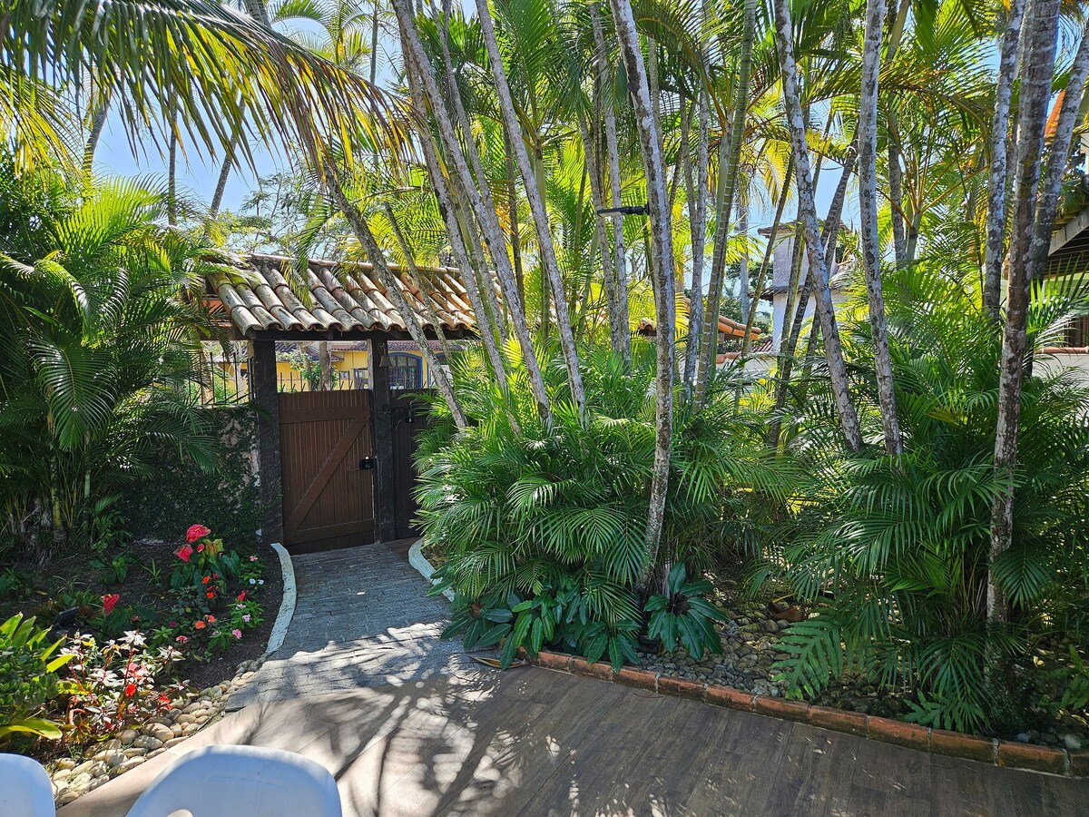 A lush garden path meanders through vibrant tropical foliage, leading to a wooden gate. The area is adorned with various plants and flowers, creating a serene atmosphere. Sunlight filters through the leaves, illuminating the natural beauty of the surroundings.