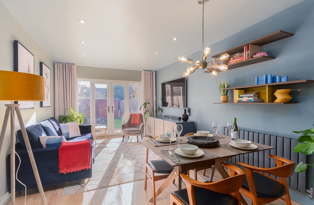 A bright living area features a modern dining table set for six, surrounded by stylish chairs. A comfortable sofa with colorful cushions and a unique floor lamp create a cozy ambiance. Large French doors lead to the garden, enhancing the natural light in the space.