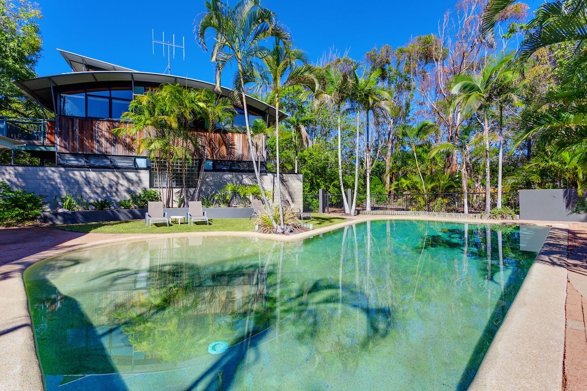 The outdoor area features a large swimming pool with beach entry, surrounded by palm trees and manicured landscaping. Comfortable seating is visible, providing spaces for relaxation under the clear blue sky. The modern structure reflects sunlight, enhancing the inviting atmosphere of the space.
