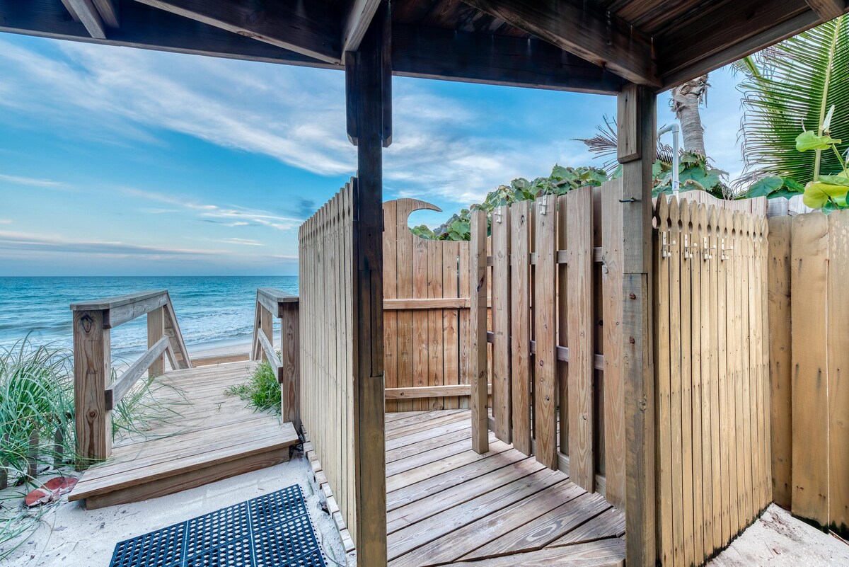 A private wooden pathway leads from the beach house to the sandy beach, surrounded by lush vegetation. A wooden gate opens to a serene coastal view, offering direct access to the tranquil ocean, where gentle waves can be seen meeting the shore.