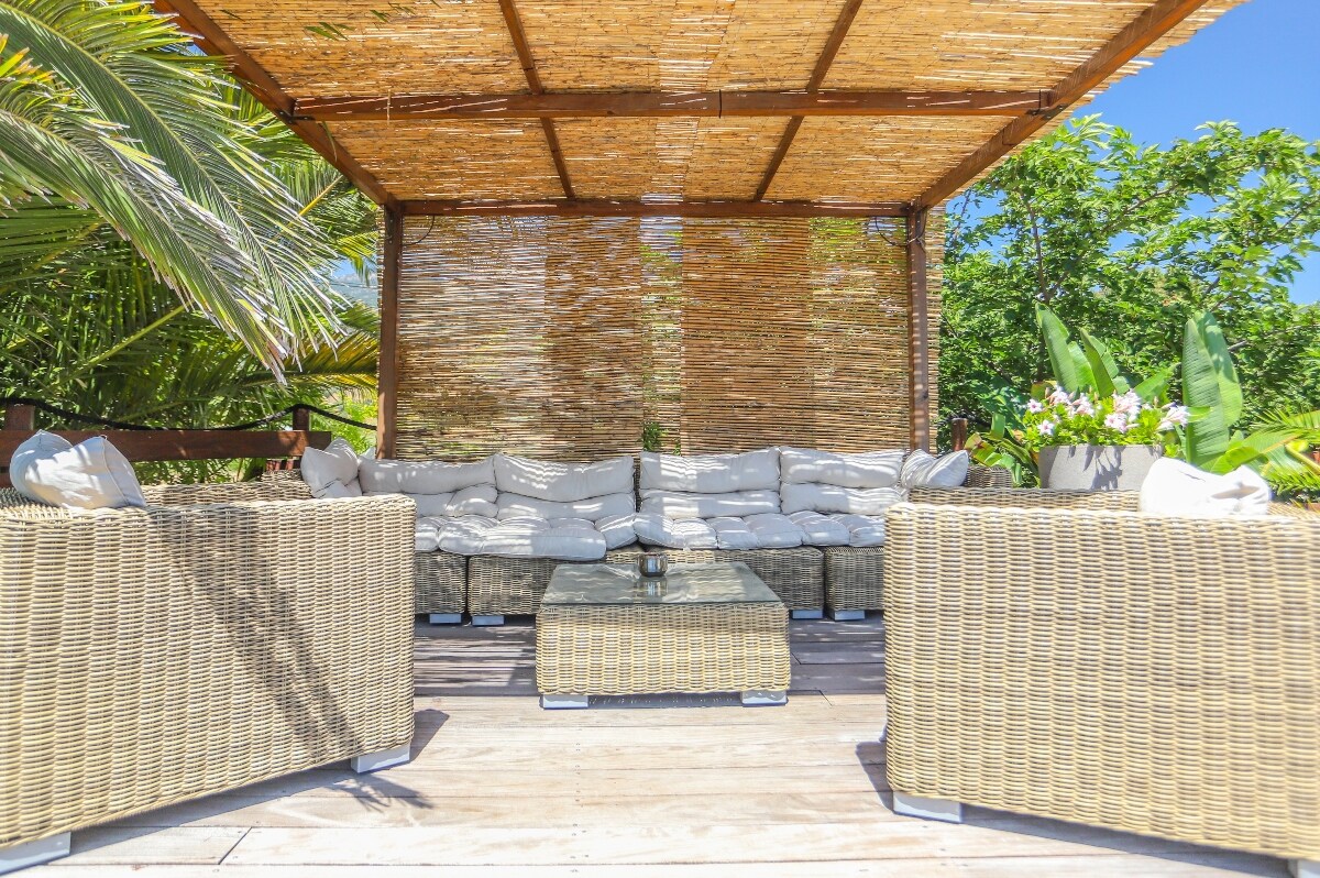 A covered terrace area features comfortable seating with light-colored cushions set around a low wooden table. Surrounding greenery and palm leaves create a natural and relaxing atmosphere, providing a shaded space for gatherings.