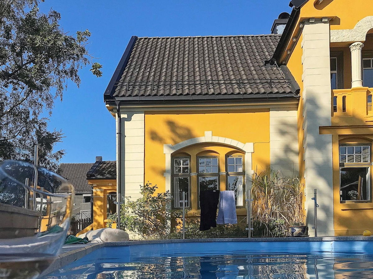 The outdoor swimming pool reflects the vibrant yellow facade of the holiday home. Nearby, windows offer glimpses into the interior, while hanging laundry adds a touch of casual domesticity. Decorative plants contribute to the welcoming environment under a clear blue sky.