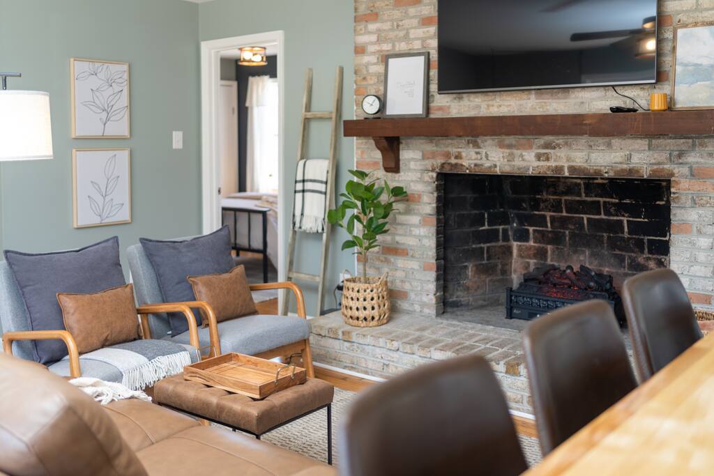 A cozy living area features two armchairs paired with a sofa, all arranged around a rustic brick fireplace. A wall-mounted television is positioned above the hearth, while light is provided by a nearby lamp. A tasteful plant adds a touch of greenery to the space.