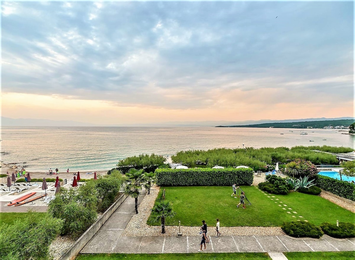 A panoramic view showcases the tranquil Adriatic Sea, framed by lush greenery and meticulously landscaped grounds. The beachfront area is visible, with guests enjoying the surroundings. Gentle waves can be seen lapping against the shore, creating a serene atmosphere at sunset.