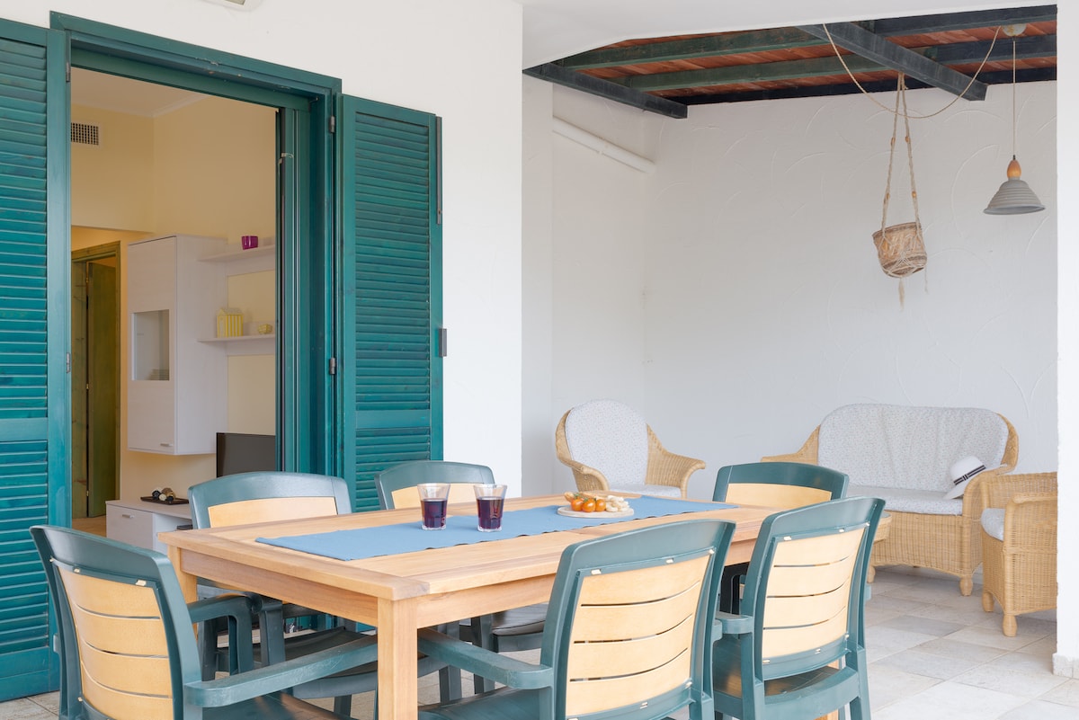 An outdoor dining area features a wooden table surrounded by six chairs, with two additional seating options nearby. Green shutters frame the entrance to an interior room, while light colors enhance the airy atmosphere. Decorative items like a hanging basket and pendant light add subtle interest.