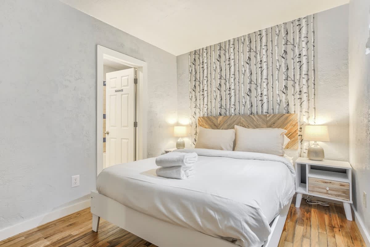 A restful bedroom features a spacious bed adorned with hotel-quality linens and neatly folded towels. Soft lighting is provided by bedside lamps, complementing the nature-inspired wallpaper. A doorway leads to the adjoining area, while wood flooring adds warmth to the overall design.