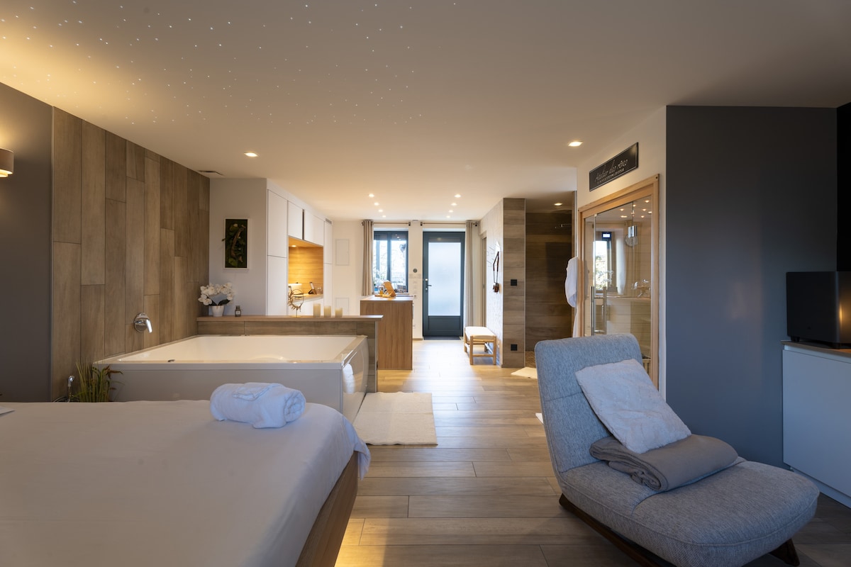 A spacious suite features a large elevated bed and a luxurious spa bathtub. Warm wood tones are complemented by modern decor. Soft lighting highlights the area, while a sauna and shower can be seen in the background. A cozy seating area invites relaxation.