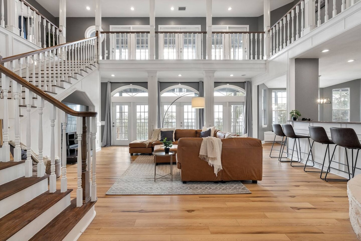 An open-concept living area displays high ceilings with large windows allowing natural light to fill the space. A spacious modular sofa is featured in the center, complemented by a decorative rug. A staircase leads to the upper level, enhancing the room's elegance.