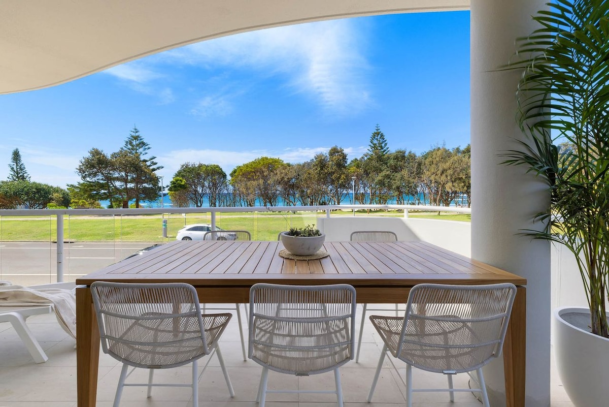 A spacious balcony features a wooden dining table surrounded by six white chairs. Lush greenery is visible in the background, along with a clear view of the beach and ocean under a blue sky.