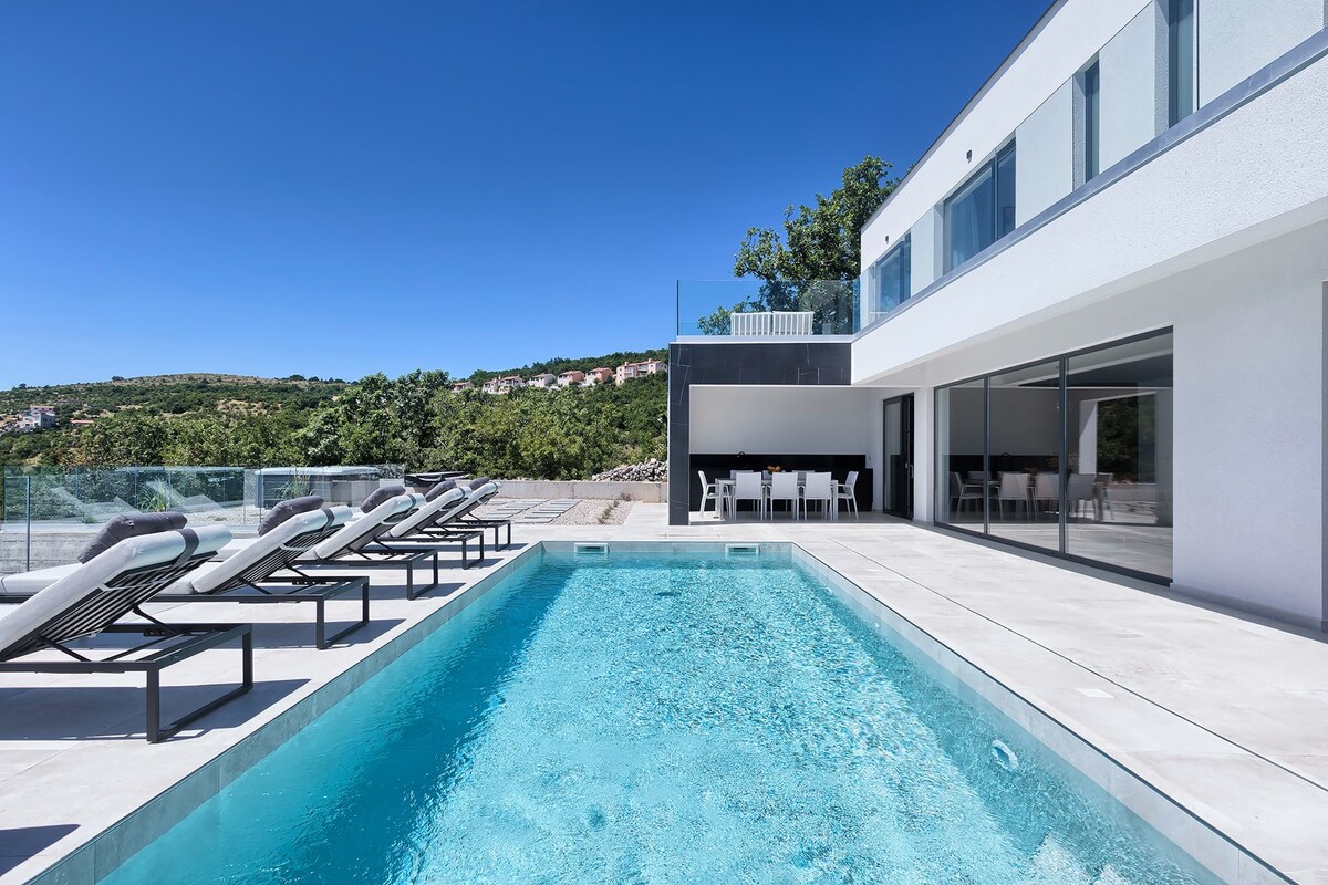 A modern outdoor area features a spacious pool surrounded by several lounge chairs. Large glass doors connect the pool area to the interior, where a dining space is visible. The clear sky and surrounding greenery create a serene backdrop.