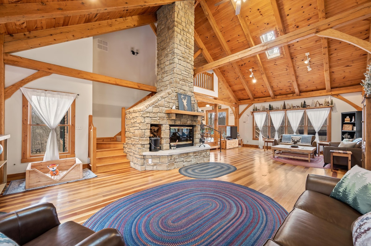 A spacious living area features a Brown County stone fireplace reaching the vaulted ceiling, surrounded by large windows draped with sheer curtains. Natural wood beams enhance the rustic charm, while plush furniture invites relaxation. A braided rug anchors the open space, providing warmth and texture.