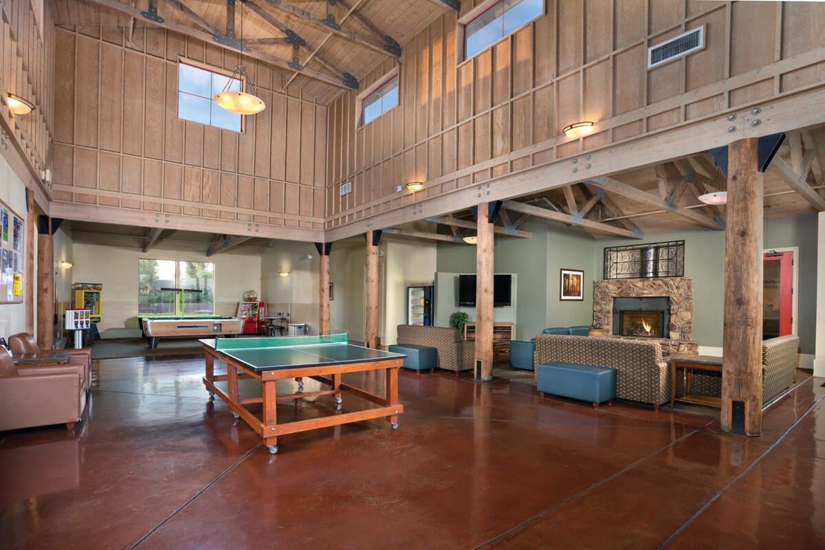 An open common area features high wooden ceilings and large windows allowing natural light to fill the space. Seating arrangements consist of leather chairs and wicker furniture surrounding a fireplace. A game table occupies the center, with additional recreational facilities visible in the background.