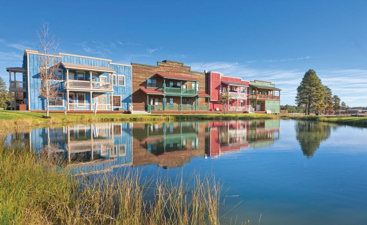 A scenic view of multi-colored, Old West-style buildings is presented along the water’s edge. Their vibrant hues are mirrored on the calm surface of the pond, surrounded by lush grass. Tall trees form a backdrop against a clear blue sky.