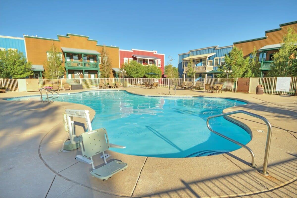 An outdoor swimming pool is surrounded by a spacious deck area, featuring several lounge chairs and tables. A pool lift is positioned at the shallow end, offering accessibility. Colorful buildings with balconies can be seen in the background, set against a clear blue sky.