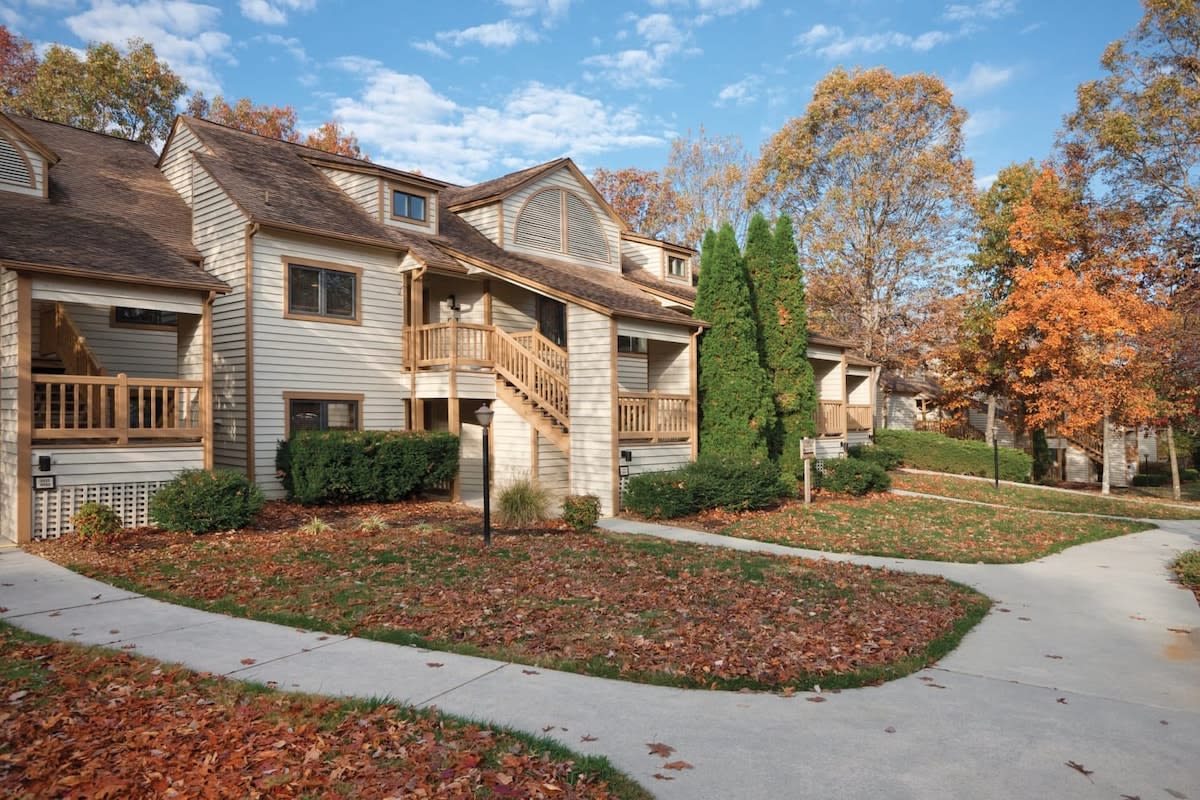 An exterior view of a two-story building is presented, framed by vibrant autumn leaves. The structure features a wooden deck with steps leading up, flanked by neatly manicured shrubs and seasonal foliage. A winding concrete pathway connects to other units in the serene environment.