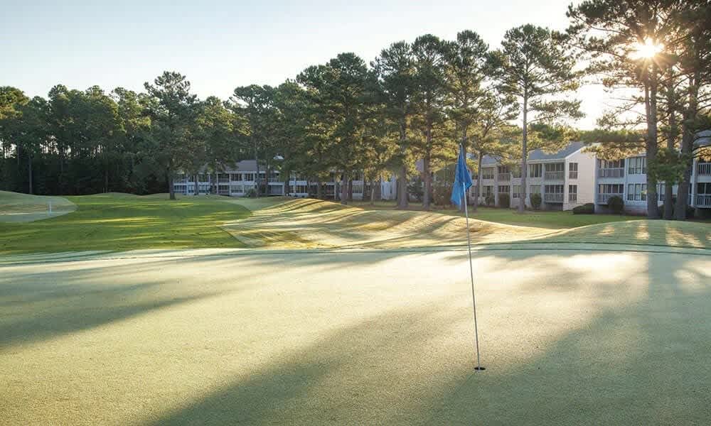 A serene golf course is seen in the early morning light, with lush green fairways and a flag gently waving on the putting green. In the background, multiple suites line the horizon, partially obscured by tall trees casting long shadows.