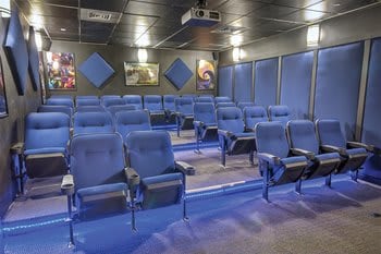 A movie theater room features rows of blue, cushioned seats arranged in a staggered formation. Wall-mounted art pieces add color, while the ceiling is equipped with projector lighting. Concealed LED lighting along the floor provides a subtle illumination.