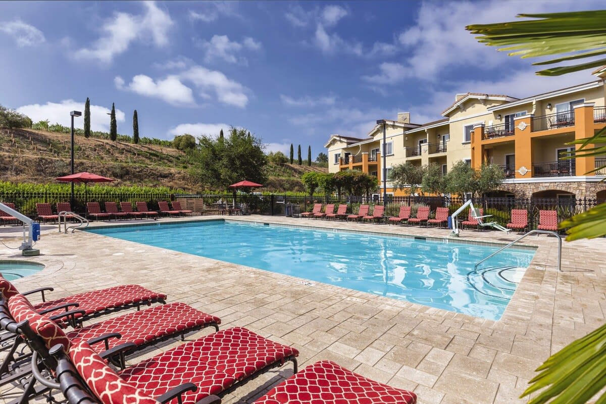 A spacious swimming pool is framed by a large stone patio with multiple lounge chairs featuring red patterns. Umbrellas provide shade around the area, while the backdrop displays a well-maintained resort building and lush greenery in the distance.