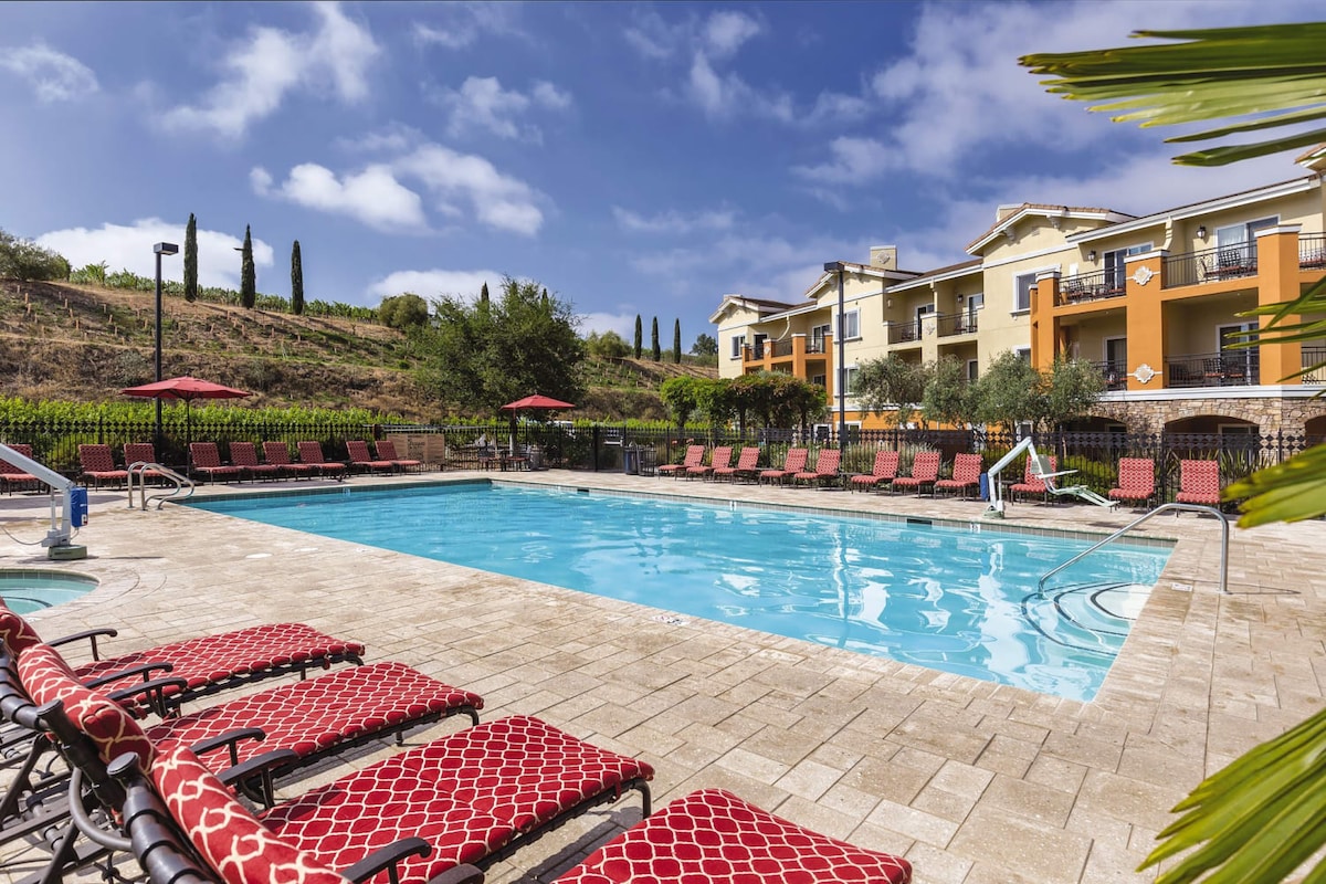 A spacious outdoor pool is surrounded by a stone deck, featuring multiple lounge chairs with red patterned cushions. Umbrellas provide shade, and the lush green landscape is visible in the background, complemented by a tastefully designed resort building.