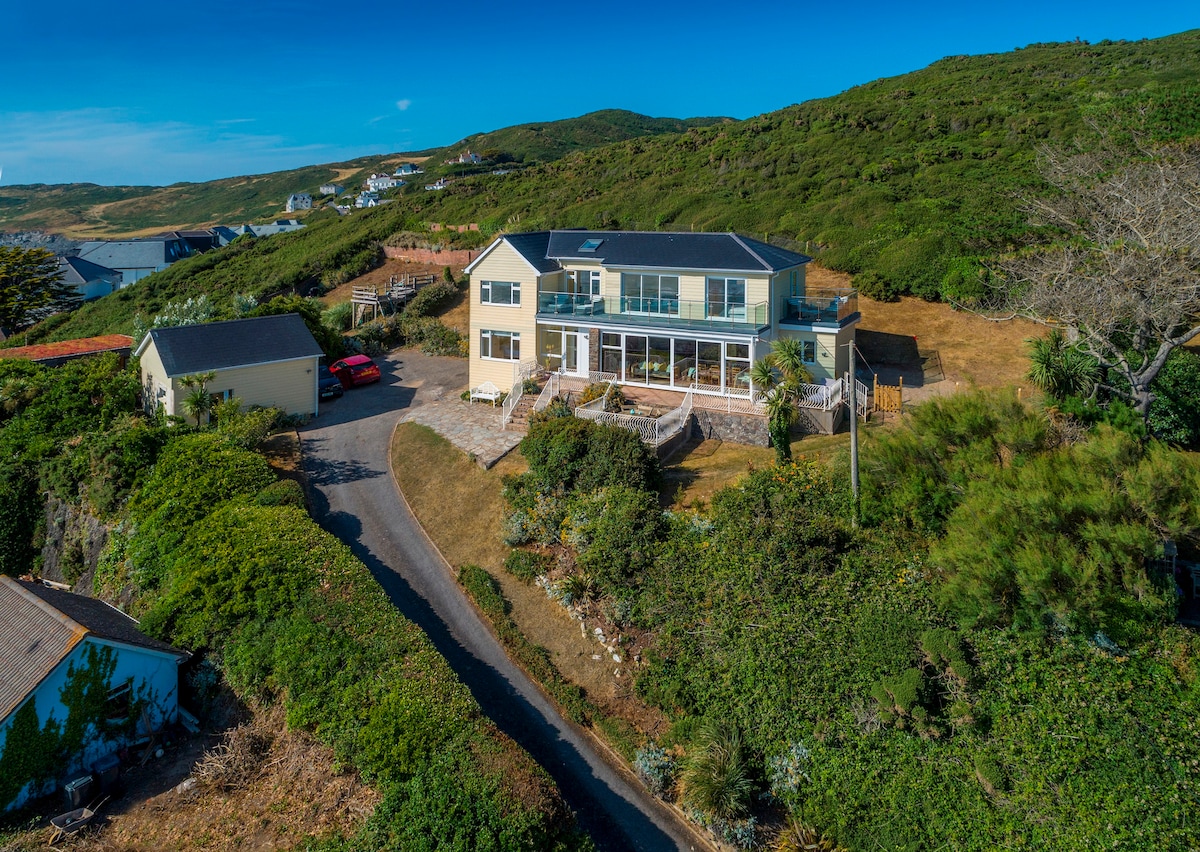 A spacious five-bedroom house is set against a backdrop of green hills. Large windows reflect panoramic sea views. The property features a landscaped garden and a winding drive leading to private parking. Outdoor seating is arranged on the terrace for enjoying the serene surroundings.