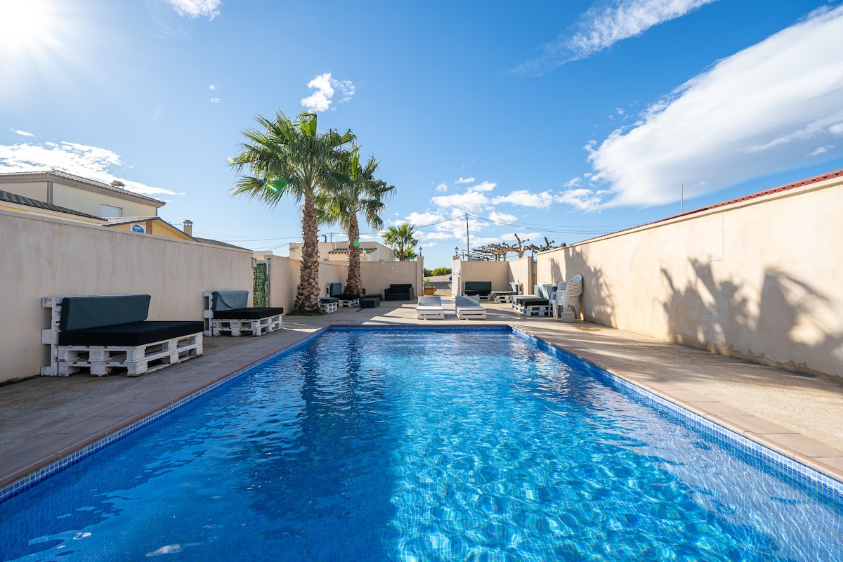 A shared outdoor pool area is visible, surrounded by palm trees and complemented by loungers made from wooden pallets. The clear blue water reflects the sunlight, while a surrounding wall provides privacy, and a blue sky with scattered clouds is seen above.