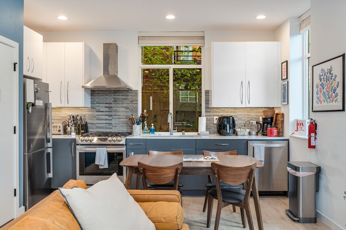 A modern kitchen features stainless steel appliances, including a gas stove and refrigerator. Light wood cabinets and a tiled backsplash complement the design. A dining table with four wooden chairs is visible, along with a cozy seating area, ensuring a functional and inviting space.