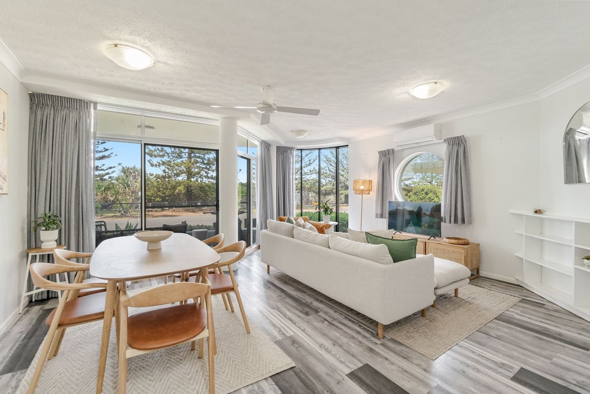 A light-filled living area features a comfortable sofa and a dining table with wooden chairs, surrounded by large windows that showcase the outdoor scenery. A ceiling fan and neutral-colored decor add to the airy ambiance.