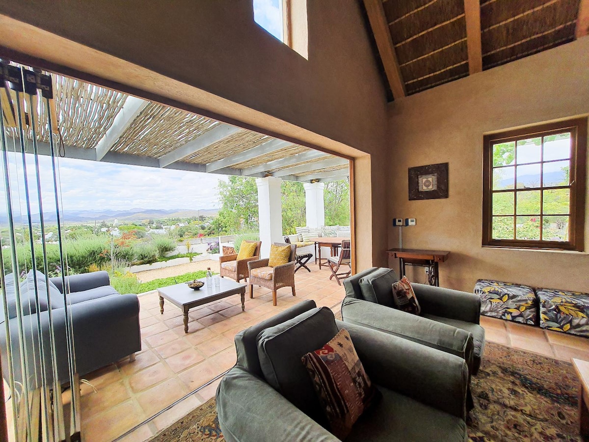An open-plan living area features glass doors that connect to a verandah. Comfortable seating arrangements are present, complemented by a coffee table. Natural light fills the space, highlighting the earthy tones of the walls and the texture of the thatched ceiling.