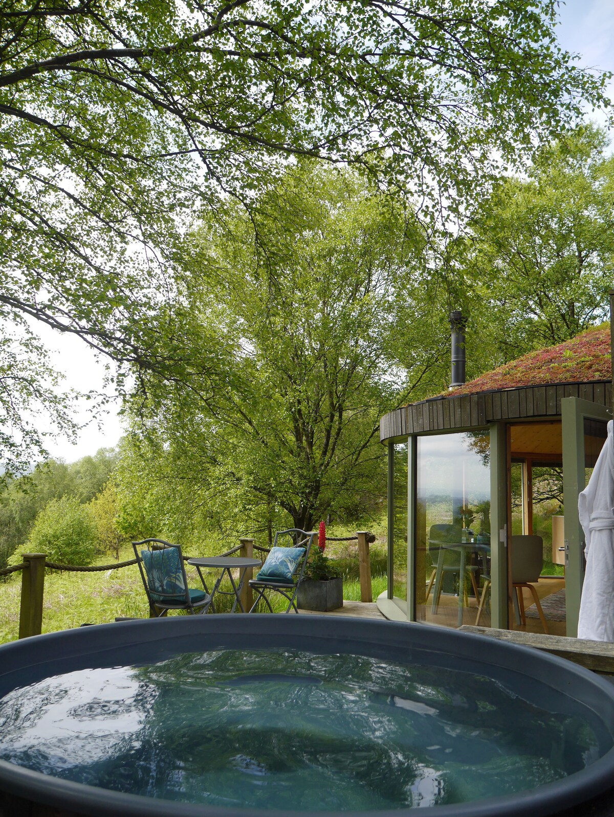 A inviting hot tub is situated on a deck surrounded by lush greenery and mature trees. Two chairs and a small table are seen nearby, offering a serene spot to relax and enjoy the peaceful countryside views.
