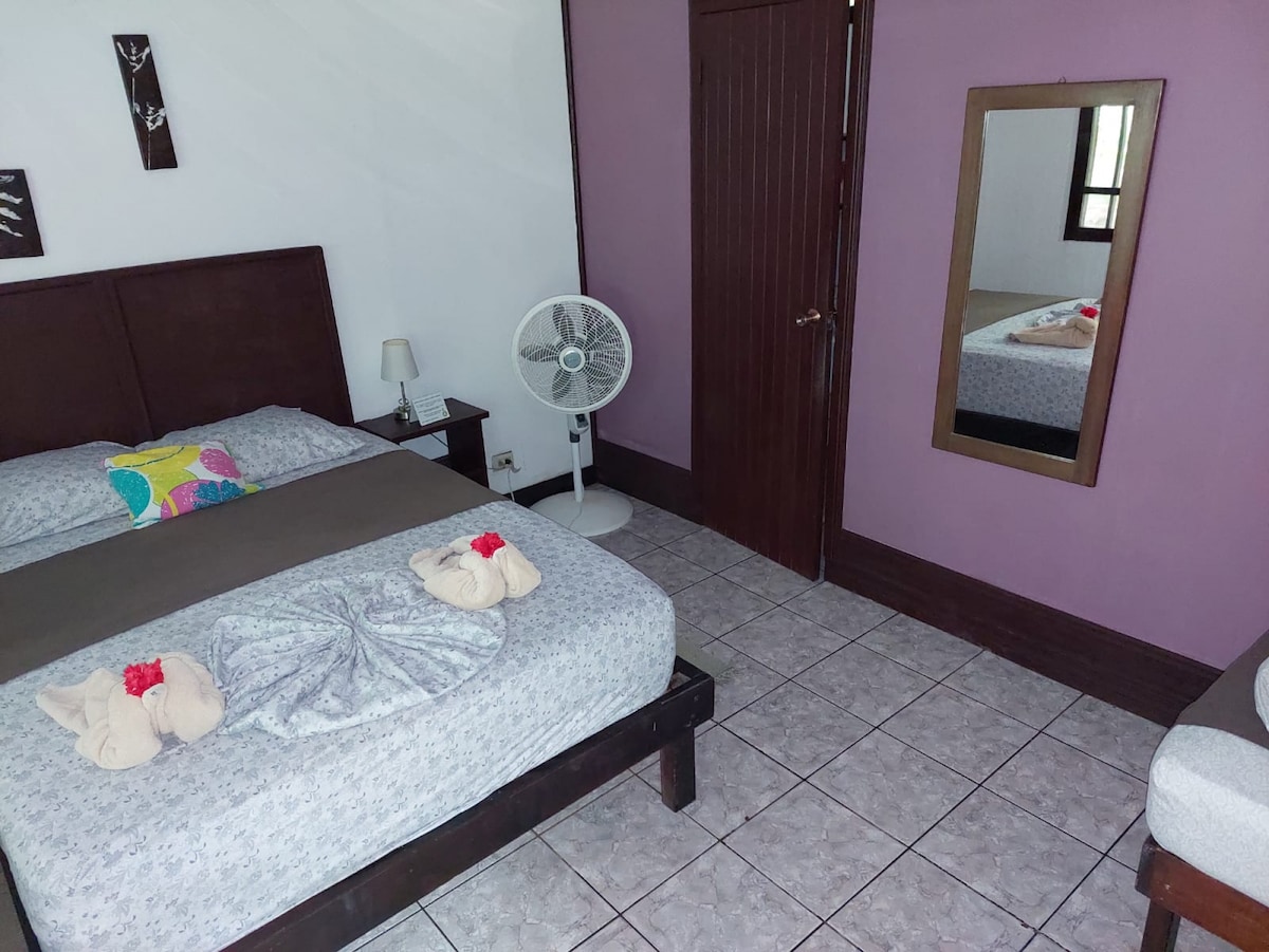 A cozy sleeping area features a double bed and a small single bed, both adorned with light bedding. A fan stands nearby, and a mirror reflects the space. Colorful decorative elements add warmth, complemented by natural light entering from a window.