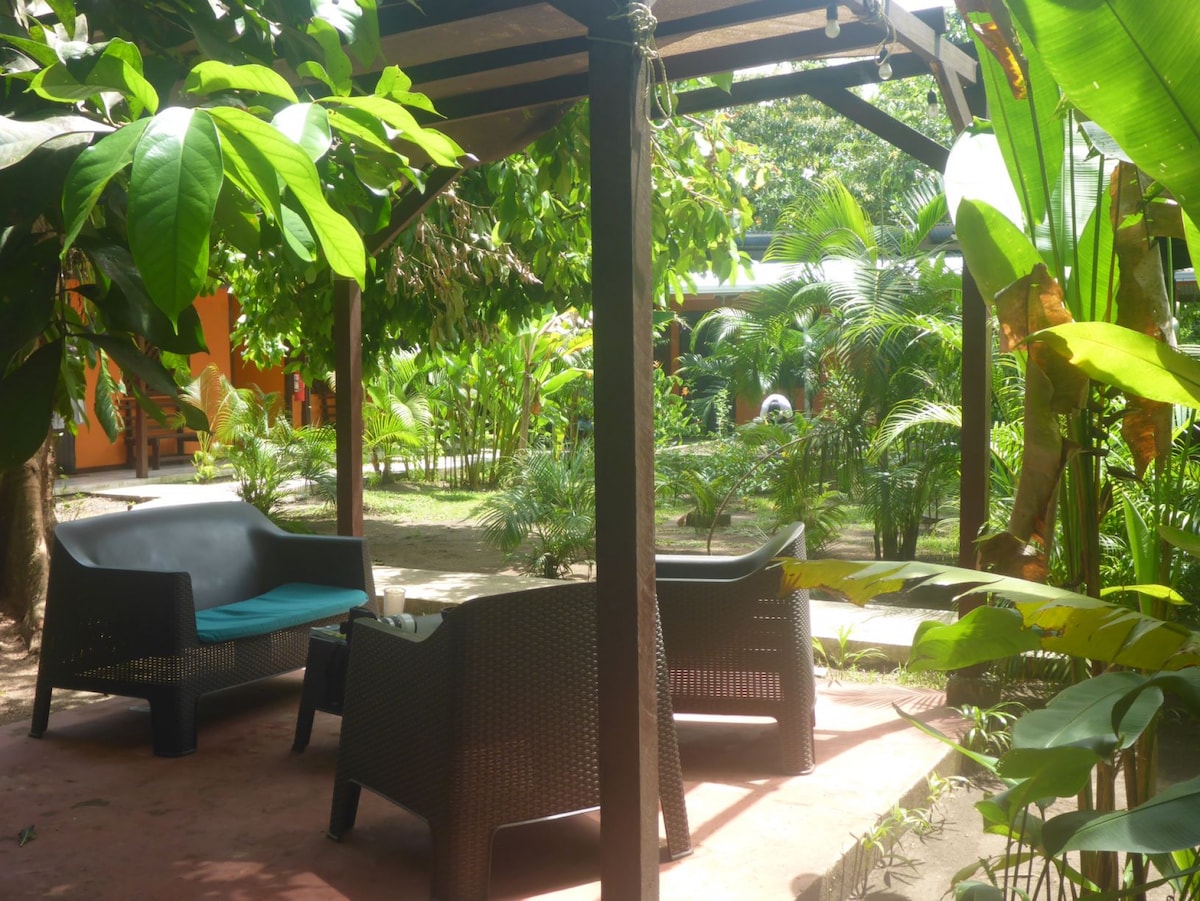 An outdoor seating area features two dark, modern chairs with teal cushions, positioned beneath a wooden pergola. Lush greenery, including palm and tropical plants, surrounds the space, providing a serene atmosphere for relaxation.