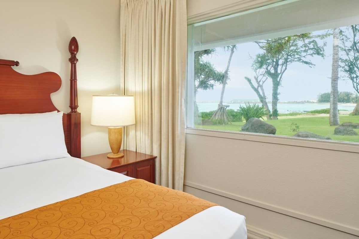 A cozy hotel room features a spacious bed with a decorative orange bed runner. Natural light enters through a large window, offering a view of lush green grass and palm trees outside, alongside Kahului Bay.