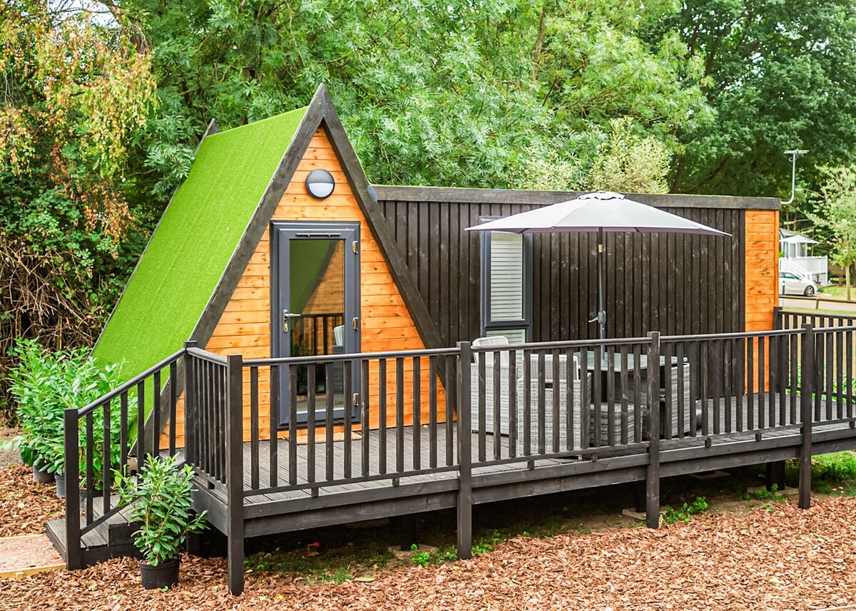 A distinct accommodation structure features a triangular green roof and warm wooden exterior. A spacious deck with seating is visible, surrounded by lush greenery. An umbrella provides shade over the seating area, enhancing the outdoor space for relaxation.