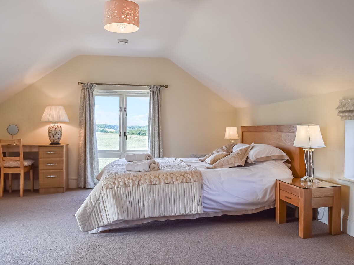 A well-lit bedroom features a spacious bed adorned with neutral bedding and decorative pillows. Two bedside lamps cast a gentle glow, while a small desk and chair are positioned nearby. Windows open to scenic views of the surrounding countryside, enhancing the soothing ambiance.