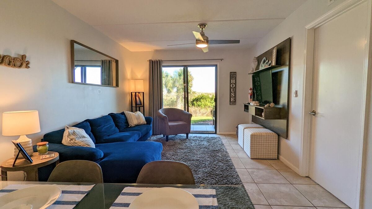 A cozy living area is showcased, featuring a blue sectional sofa and a comfortable armchair. A ceiling fan provides airflow, while a large window invites natural light and offers views of the outdoor greenery. A decorative mirror and stylish shelving accent the space.