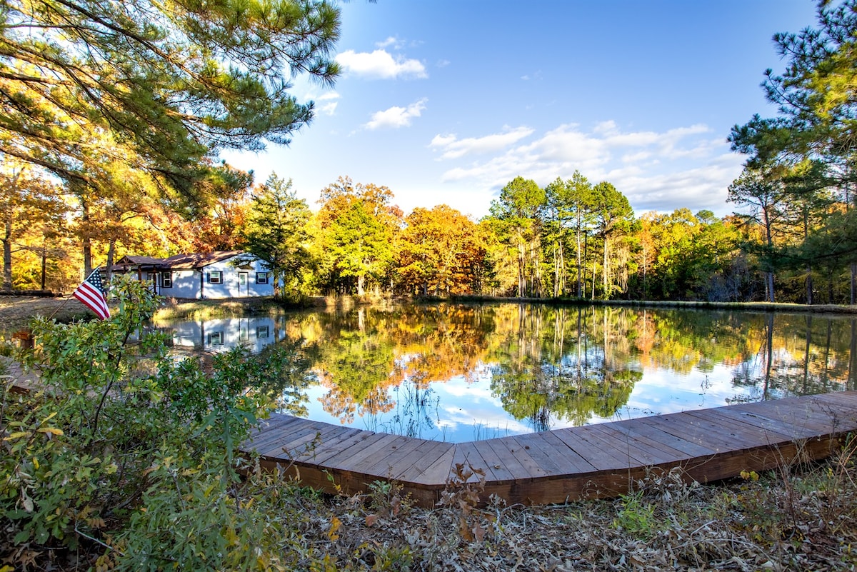 A serene pond reflects vibrant autumn colors from surrounding trees, with a wooden deck leading to the water's edge. An American flag subtly flutters in the breeze, contributing to the tranquil setting of Birch Cabin.