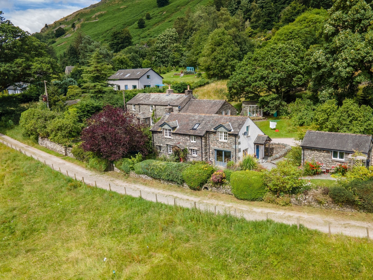 A charming stone cottage is nestled amidst lush greenery, characterized by a winding path leading to it. Surrounding trees and shrubs enhance the natural setting, while the structure itself features multiple sections with a rustic appeal. The backdrop consists of rolling hills, providing a serene rural atmosphere.
