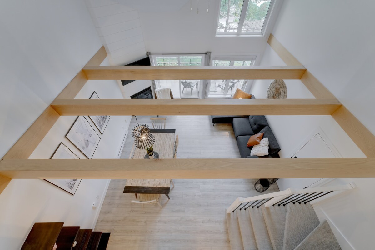 An elevated view of the living area showcases a spacious layout with large windows allowing natural light. A modern couch is positioned adjacent to a dining table, with decorative elements enhancing the space. The open design connects the upper and lower levels seamlessly.