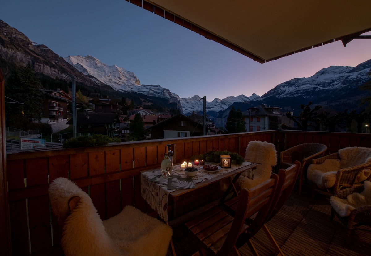 An outdoor balcony area features wooden flooring and a rustic table set for dining. Soft blankets drape over chairs, while candles provide warm light. In the background, the majestic snow-capped mountains are illuminated by the soft glow of dusk.