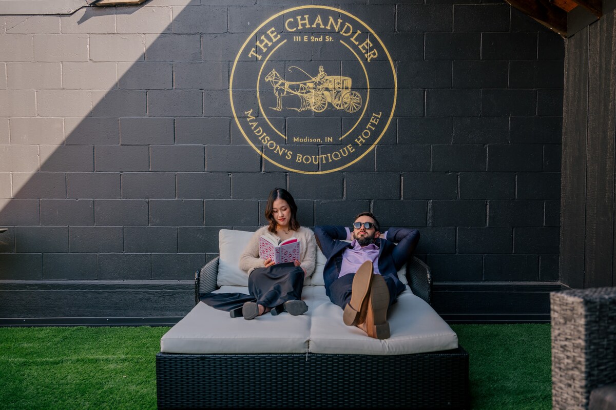A cozy outdoor seating area features a spacious, cushioned lounge sofa positioned against a dark wall. The Chandler logo is displayed prominently above. Soft green grass complements the setting, while two guests relax, one reading a book and the other reclining comfortably.
