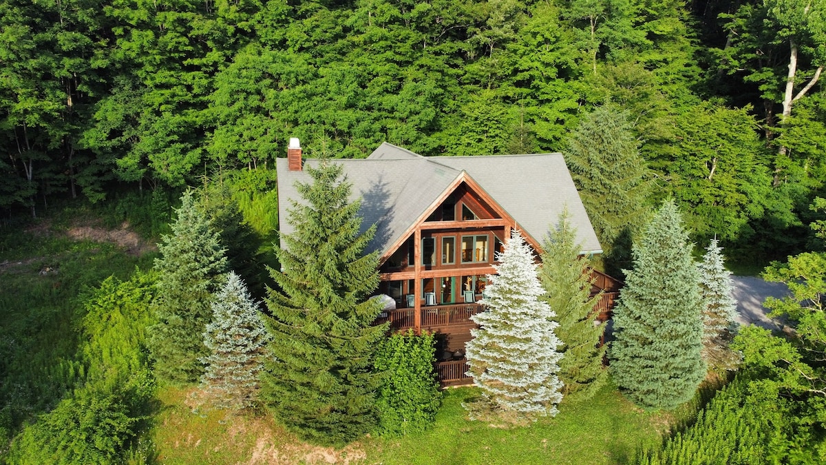 A spacious chalet is nestled among dense greenery, surrounded by tall trees that provide both privacy and natural beauty. The structure features a combination of wood and stone elements, with large windows that invite ample natural light, creating a serene atmosphere.
