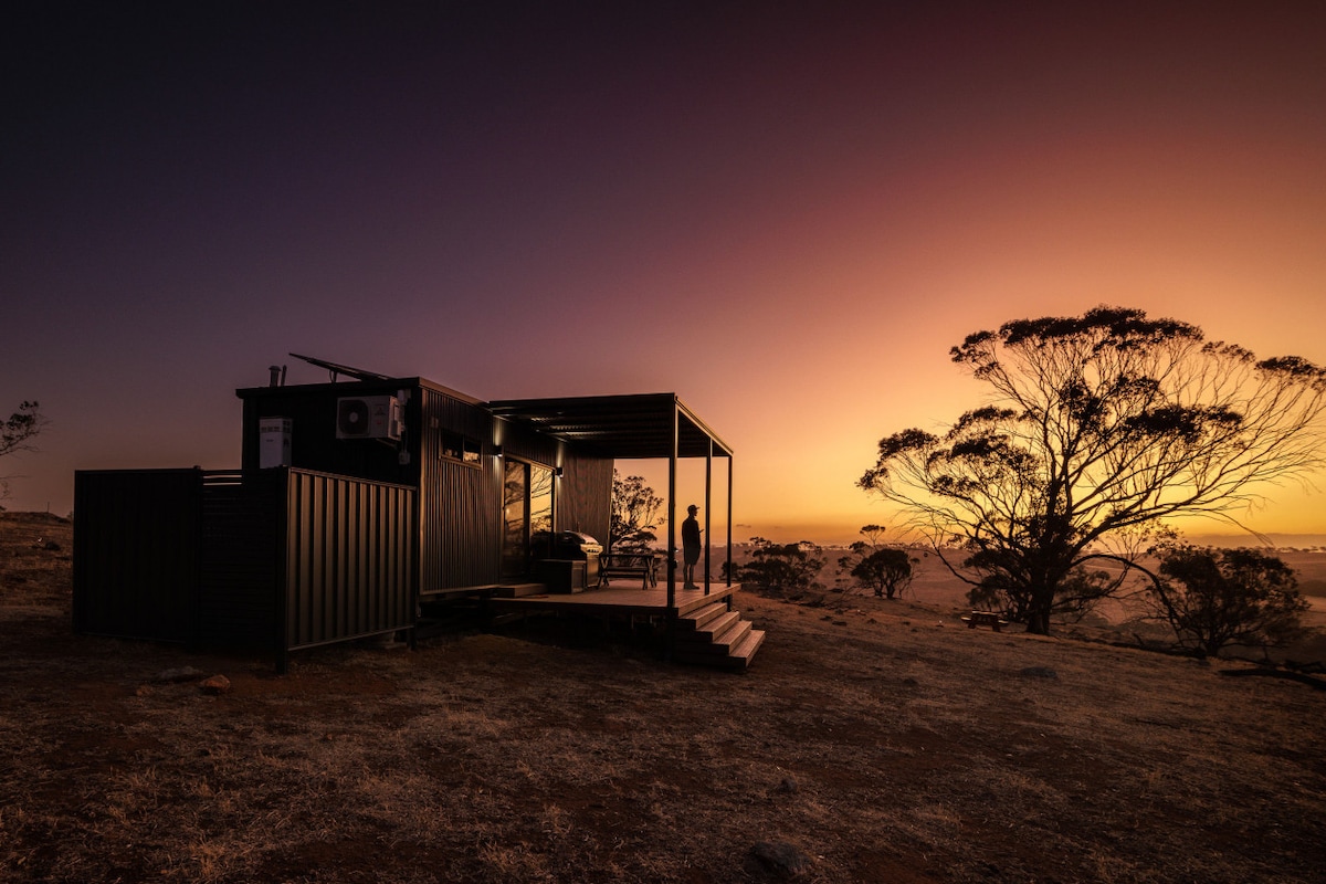 The cabin is positioned on open land with a deck that provides a view of the horizon. Soft lighting illuminates the exterior, while trees can be seen silhouetted against a colorful sunset sky.
