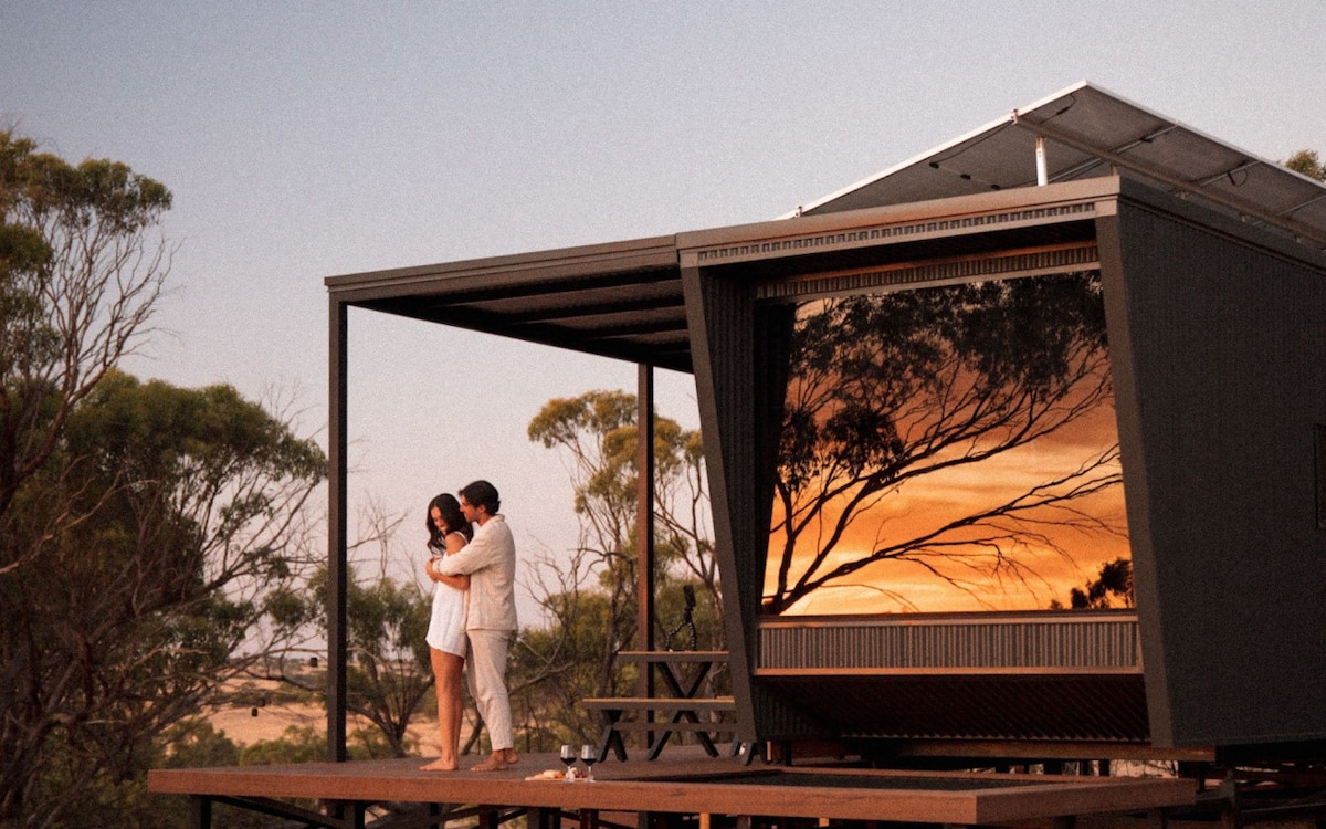 The tiny cabin is surrounded by trees, showcasing a striking sunset reflected in its large window. A wooden deck features a picnic table, inviting relaxation. Soft hues of orange and purple fill the sky, creating a serene backdrop for the cabin's modern design.