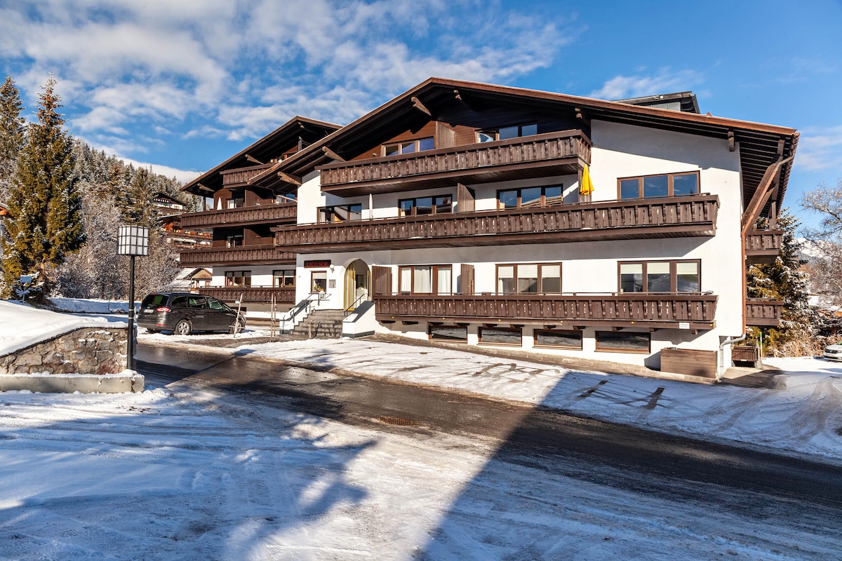 A three-story building features a wooden façade with balconies overlooking the surroundings. The property is set against a snowy landscape, with pine trees in the background. A partially cleared driveway leads to the entrance, and parking spaces are visible in the foreground.