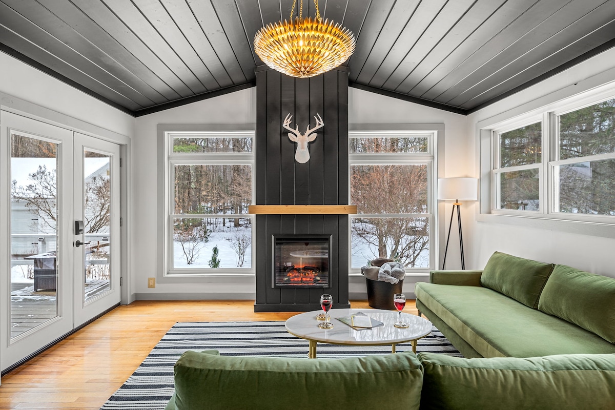 A cozy sitting area is presented with a green sectional sofa and a round coffee table. A modern chandelier hangs above, and a faux deer head is mounted on the dark wood accent wall. Large windows invite natural light, revealing a snowy landscape outside.